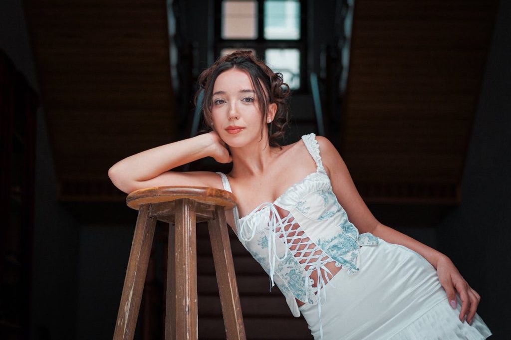 A woman in a white and blue dress poses elegantly indoors, leaning on a wooden stool.