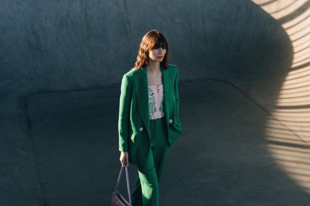 Fashionable woman in green blazer and pants holding a handbag in a minimalist outdoor setting.